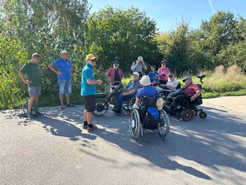Menschen stehen im Schatten eines Baumes. Einige Personen sitzen im Rollstuhl.
