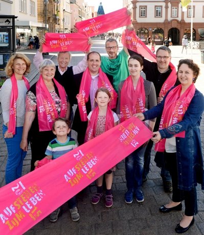Gruppe von jungen und älteren Menschen stehen auf der Speyerer Hauptstraße und halten die Magenta-farbenen Teilnehmerschals zum Ökumenischen Kirchentag hoch.